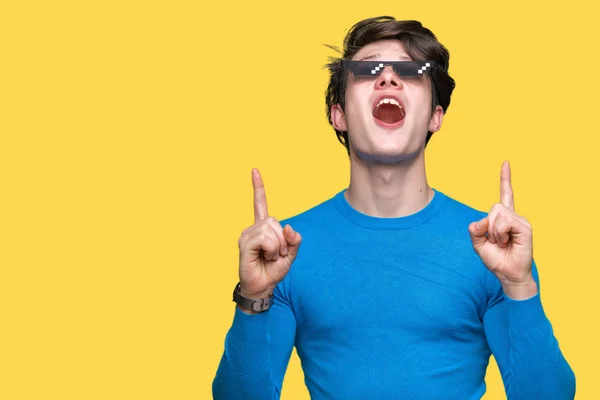 Young man wearing funny thug life glasses over isolated background amazed and surprised looking up and pointing with fingers and raised arms.