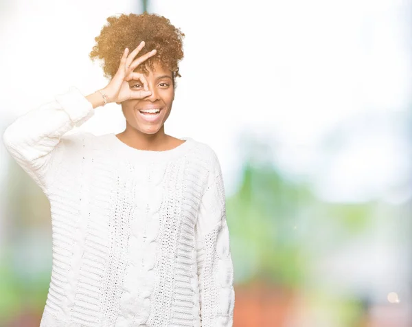 Beautiful young african american woman wearing winter sweater over isolated background doing ok gesture with hand smiling, eye looking through fingers with happy face.