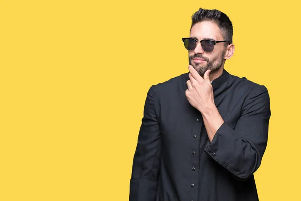 Young Christian priest wearing sunglasses over isolated background with hand on chin thinking about question, pensive expression. Smiling with thoughtful face. Doubt concept.