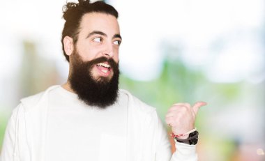 Uzun saç ve sakal seyir ve başparmak yanında yukarıyı mutlu yüz ile gülümseyen sportif sweatshirt giyen genç adamla.