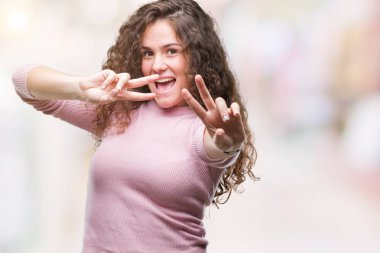 Güzel esmer kıvırcık saçlı Genç kız zafer işareti yapıyor parmak gösterilen kamera seyir gülümseyen izole arka plan üzerinde pembe kazak giyiyor. İki numaralı.