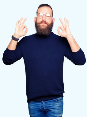 Gözlük ve balıkçı yaka kazak giyen genç sarışın adam rahatla ve gülümseyen gözlerle kapalı parmak jestle meditasyon yapıyor. Yoga kavramı.