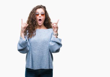 Güzel esmer kıvırcık saçlı Genç kız izole arka plan üzerinde güneş gözlüğü takıyor hayret ve yukarıya bakıp parmakları ile işaret şaşırttı ve silah kaldırdı.