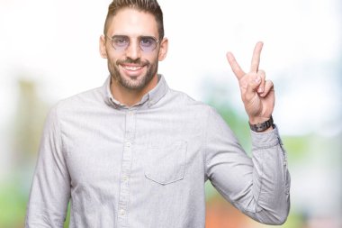 Genç yakışıklı adam mutlu yüz zafer işareti yaparken kameraya göz kırpıyor ile gülümseyen izole arka plan üzerinde güneş gözlüğü takıyor. İki numaralı.
