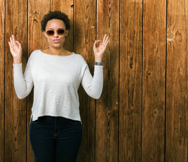 Güzel genç Afro-Amerikan kadın giyiyor güneş gözlüğü izole arka plan üzerinde dinlenmek ve gülümseyen gözlerle kapalı parmak jestle meditasyon yapıyor. Yoga kavramı.