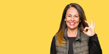 Güzel orta yaş giyen kış yelek el ve parmaklar ok işaretiyle yapıyor olumlu gülümseyen kadın. Başarılı ifade.