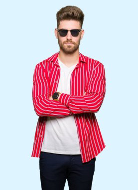 Çapraz kolları olan yüzündeki güneş gözlüğü şüpheci ve sinir, onaylamayan ifade giyen genç yakışıklı adam. Negatif kişi.