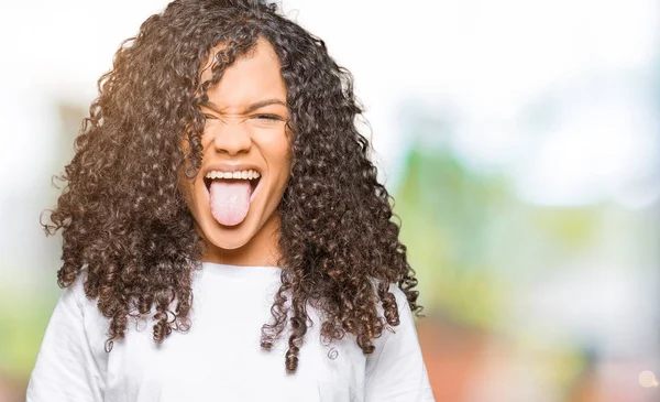 Young beautiful woman with curly hair wearing white t-shirt sticking tongue out happy with funny expression. Emotion concept.