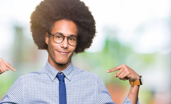 Young african american business man with afro hair wearing glasses looking confident with smile on face, pointing oneself with fingers proud and happy.