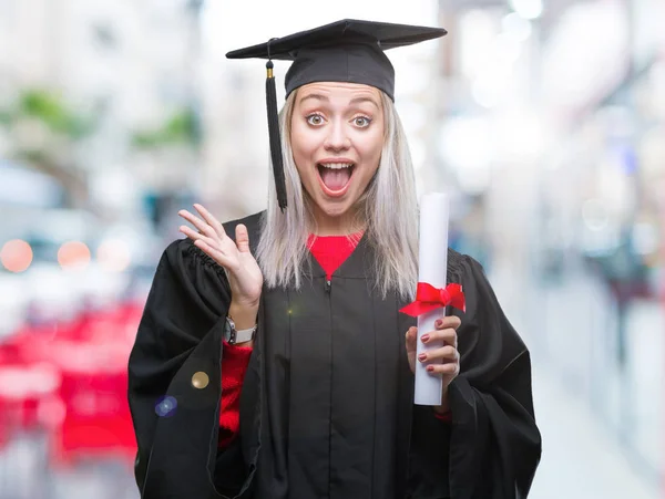 年轻的金发女子穿着研究生制服持有学位在孤立的背景下非常高兴和兴奋 优胜者表达庆祝胜利尖叫着大微笑和举手 — 图库照片