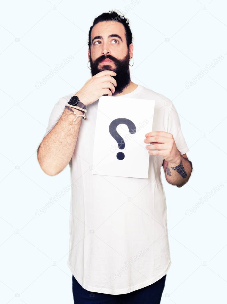 Young man with long hair and bear holding a paper with question mark serious face thinking about question, very confused idea