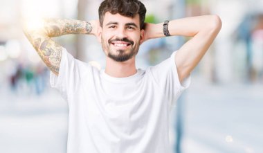 Beyaz t-shirt üzerinde izole giyen genç yakışıklı adam arka plan dinlenirken ve germe kollar ve eller baş ve boyun arkasında ile mutlu gülümseyerek