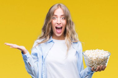 Kocaman bir gülümseme ile çığlık zaferini kutluyor ve ellerini kaldırdı izole arka plan çok mutlu ve heyecanlı, kazanan ifade üzerinde patlamış mısır yemek güzel sarışın genç kadın