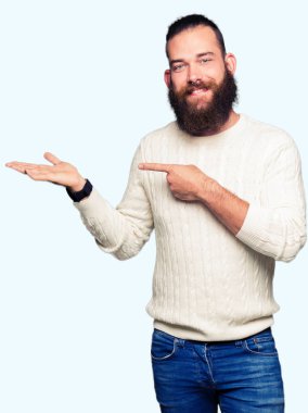 Genç hipster adam giyen kış kazak hayret ve el ile sunulması ve parmak ile işaret ederken kameraya gülümseyen.