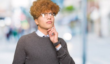 Genç yakışıklı iş adamı afro saçlı gözlüklü el ile çene düşünme hakkında soru, dalgın ifade üzerine. Düşünceli yüzüyle gülümsüyor. Şüphe kavramı.