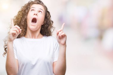 Güzel esmer kıvırcık saçlı Genç kız izole arka plan üzerinde rahat t-shirt giyen hayret ve yukarıya bakıp parmakları ile işaret şaşırttı ve silah kaldırdı.