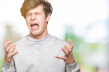 Üzerinde sweatshirt giyen genç yakışıklı sportif adam arka plan deli izole ve deli bağırarak ve agresif ifade ve silah kaldırdı bağırıyor. Hayal kırıklığı kavramı.