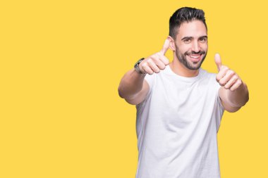 El ile pozitif jest yapıyor onaylama izole arka plan üzerinde rahat beyaz t-shirt giyen genç adam başparmak gülümseyen ve başarı için mutlu. Kamera, kazanan jest arıyorsunuz.