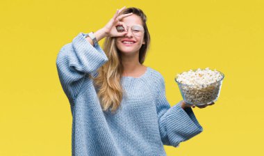 Genç güzel sarışın kadın mutlu yüz el parmaklarının arasından seyir göz üzerinde ok işaretiyle yapıyor gülümseyen izole arka plan üzerinde patlamış mısır yemek