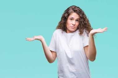 Güzel esmer kıvırcık saçlı Genç kız üzerinde rahat t-shirt giyen arka plan clueless ve şaşkın bir ifadeyle kollar ve eller kaldırdı izole. Şüphe kavramı.