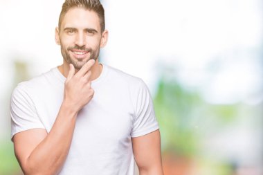 Emin çapraz kollar ve çeneme kaldırdı el ile gülümseme ile kameraya bakarak izole arka plan üzerinde rahat beyaz t-shirt giyen genç adam. Pozitif düşünce.