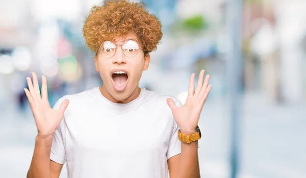 Young handsome man with afro hair wearing glasses celebrating crazy and amazed for success with arms raised and open eyes screaming excited. Winner concept