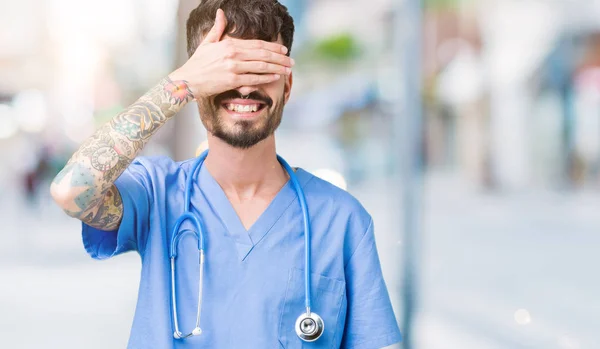 Young handsome nurse man wearing surgeon uniform over isolated background smiling and laughing with hand on face covering eyes for surprise. Blind concept.