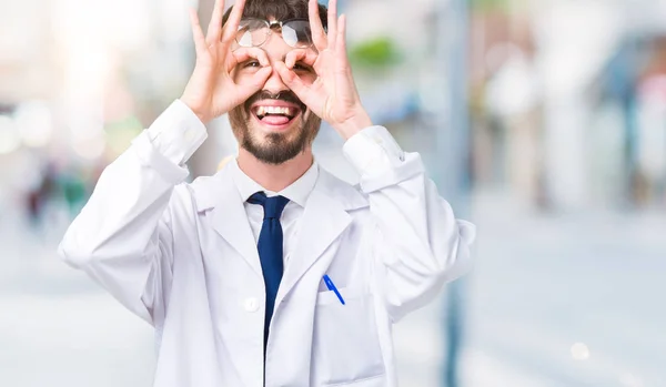 若いプロの科学者男は舌を出して指を通して見る目をこだわりの双眼鏡のような ジェスチャーを行う分離の背景に白のコートを着てします クレイジー式 — ストック写真