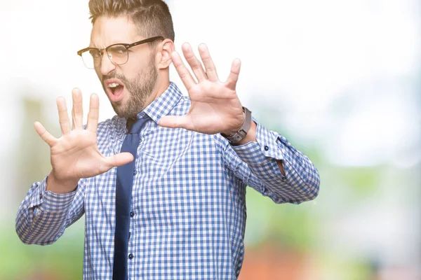 Young business man wearing glasses over isolated background afraid and terrified with fear expression stop gesture with hands, shouting in shock. Panic concept.
