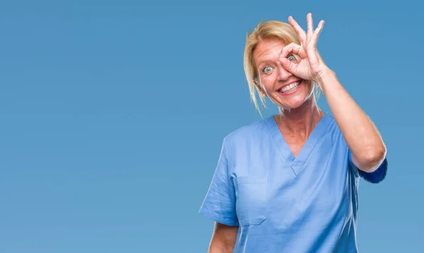 中年金发女子穿医生护士制服在孤立的背景做 手势与手微笑 眼睛看通过手指与幸福的脸 — 图库照片