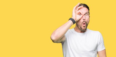 Yakışıklı adam ok jest yapıyor casual beyaz t-shirt giyen sürpriz yüz, göz parmaklarının arasından seyir ile şok. İmansız ifade.