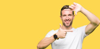 Yakışıklı adam elleriyle yapım çerçeve ve parmakları ile mutlu yüz gülümseyen casual beyaz t-shirt giyiyor. Yaratıcılık ve Fotoğrafçılık kavramı.