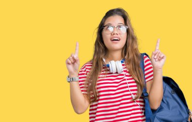 Kulaklıklar ve sırt çantası izole arka plan üzerinde giyen genç güzel esmer öğrenci kadın hayrete ve yukarıya bakıp parmakları ile işaret şaşırttı ve silah kaldırdı.