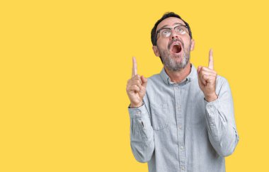 İzole arka plan üzerinde gözlük takan yakışıklı orta yaş zarif komuta sizde hayrete ve yukarıya bakıyor ve parmakları ile işaret ve silah kaldırdı şaşırttı.