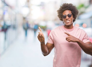 Gülümseyen ve iki elleri ve parmakları ile tarafına işaret kamera bakarak izole arka plan üzerinde güneş gözlüğü takıyor Afro Amerikalı adam.