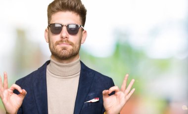 Genç yakışıklı zarif adam giyen güneş gözlüğü rahatla ve gülümseyen gözlerle kapalı parmak jestle meditasyon yapıyor. Yoga kavramı.