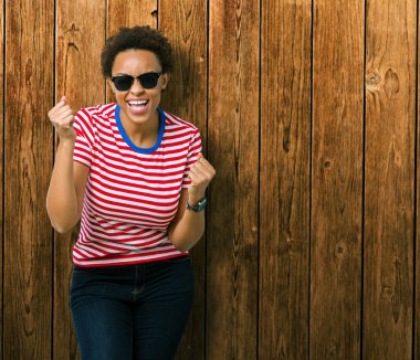 Güzel genç Afro-Amerikan kadın üzerinde güneş gözlüğü takıyor arka plan çok mutlu ve heyecanlı kaldırdı, silah ile kazanan jest yapıyor gülümseyen ve başarı için çığlık izole. Kutlama kavramı.