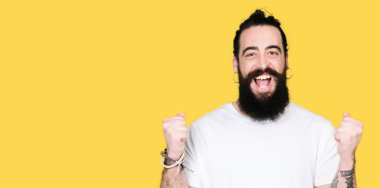 Uzun saç ve sakal silah ile şaşırmış ve hayret için başarı kutluyor casual beyaz t-shirt giyen genç hipster adamla kaldırdı ve gözleri açık. Kazanan kavramı.