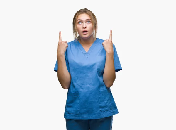 若い白人の看護師女性隔離された背景の上の外科医の制服を着てびっくりし 驚き見て 指で指して 腕を上げた — ストック写真