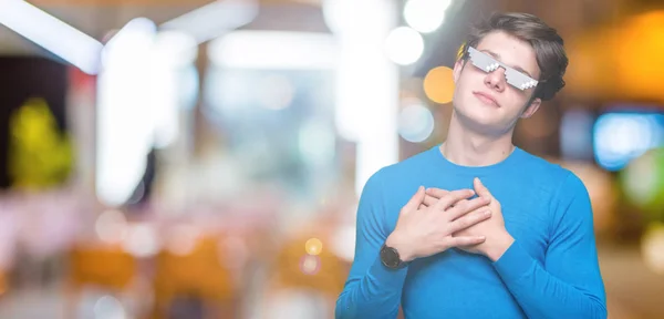 Young man wearing funny thug life glasses over isolated background smiling with hands on chest with closed eyes and grateful gesture on face. Health concept.