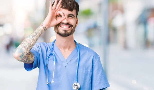 Young handsome nurse man wearing surgeon uniform over isolated background doing ok gesture with hand smiling, eye looking through fingers with happy face.