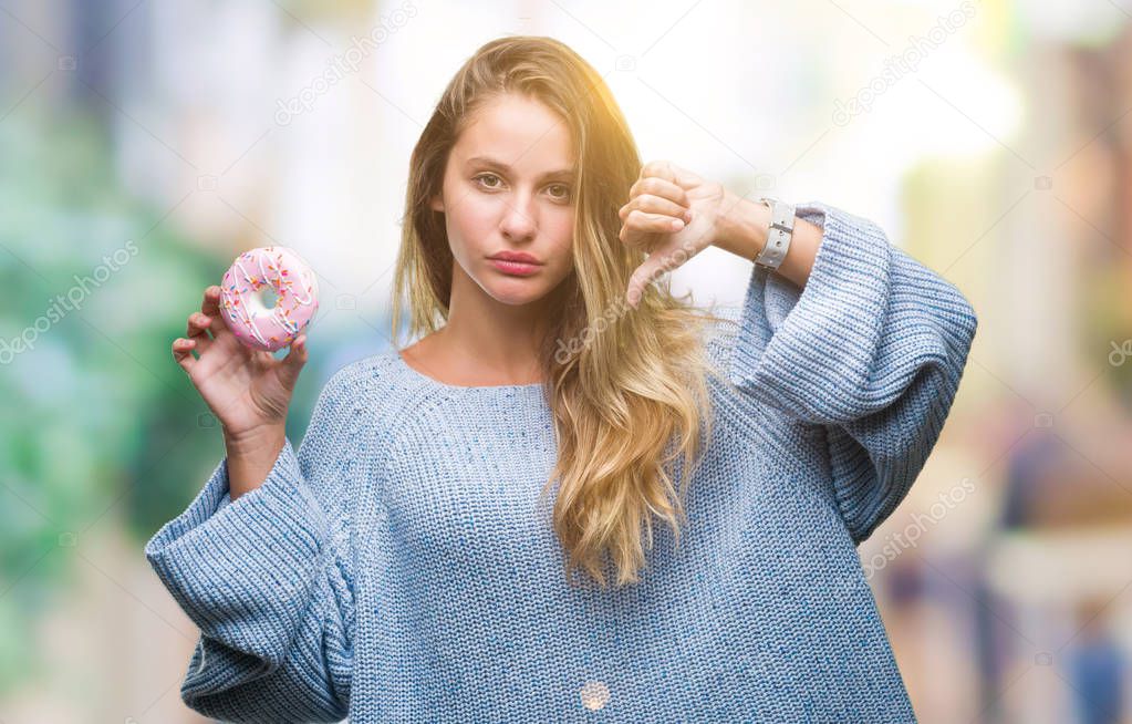 Young beautiful blonde woman eating sweet donut over isolated background with angry face, negative sign showing dislike with thumbs down, rejection concept
