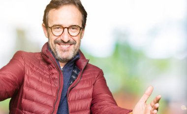 Gözlük ve kamera ile gülümseyen bakarak kış ceket giyen orta yaş yakışıklı adam silah sarılmak için açın. Neşeli ifade kucaklayan mutluluk.