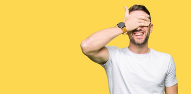 Yakışıklı adam gülümsüyor ve gözleri için sürpriz kapsayan yüz el ile gülüyor casual beyaz t-shirt giyiyor. Kör kavramı.