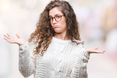 Güzel esmer kıvırcık saçlı Genç kız üzerinde kış kazak giyiyor arka plan clueless ve şaşkın bir ifadeyle kollar ve eller kaldırdı izole. Şüphe kavramı.