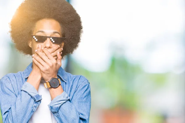 Young african american man with afro hair wearing thug life glasses shocked covering mouth with hands for mistake. Secret concept.