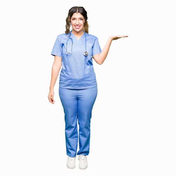 陽気な提示とカメラを見て手の手のひらで指している笑みを浮かべて医療制服を着て若い大人医師女性 — ストック写真