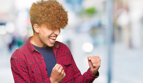 Young handsome student man with afro hair wearing a jacket very happy and excited doing winner gesture with arms raised, smiling and screaming for success. Celebration concept.