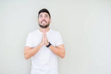 Yalvarıyor ve umut ifade yüzünde çok duygusal ve endişe ile birlikte elleriyle dua izole arka plan üzerinde rahat beyaz t-shirt giyen genç yakışıklı adam. Bağışlanmak. Din kavramı.