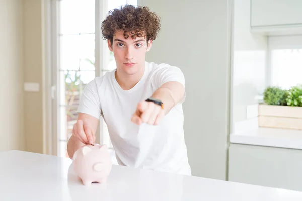 Молодий Чоловік Вкладає Гроші Використовуючи Скарбничку Вдома Вказуючи Пальцем Камеру — стокове фото